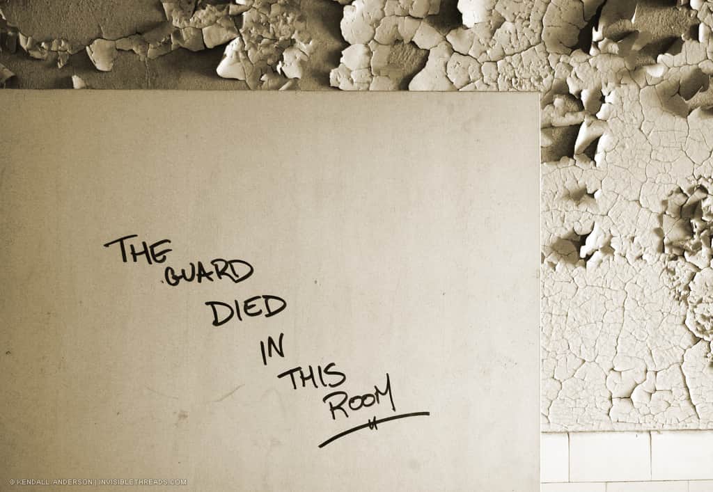A clean white door rests against a wall with peeling paint. In marker, someone has written on the door 'The Guard Died In This Room'.