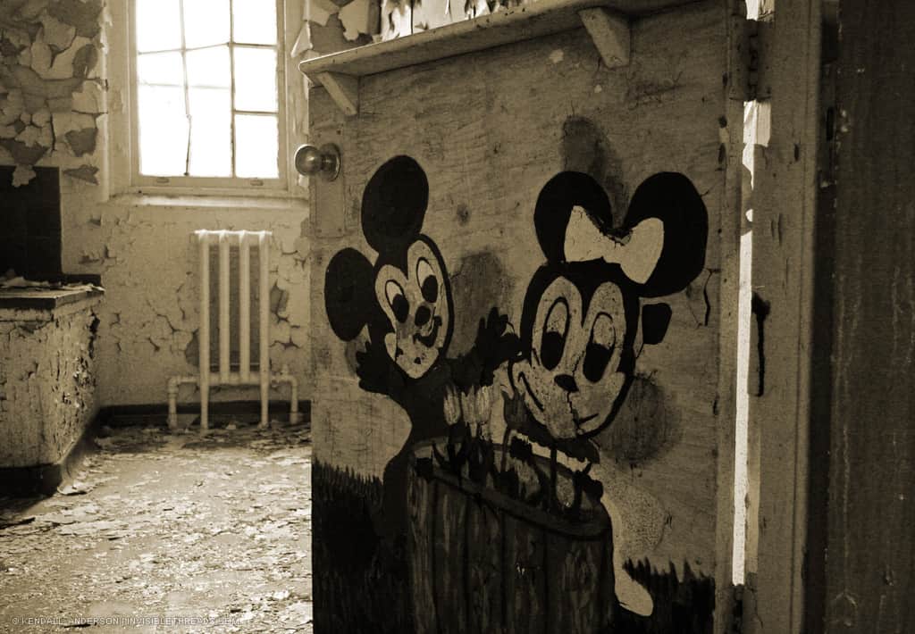 A door with painted Mickey and Minnie Mouse characters leads into an abandoned room with debris and peeling paint.