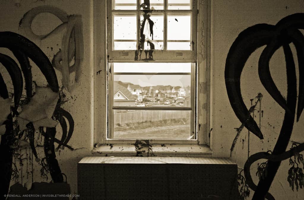 Inside an abandoned building, an interior wall with graffiti frames a window. Through the window one can see the houses of a new subdivision adjacent to the abandoned building.