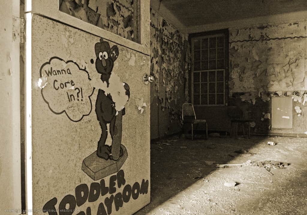 A door with a painted Elmo character leads into an abandoned room with debris and decay, and a single chair in the corner.