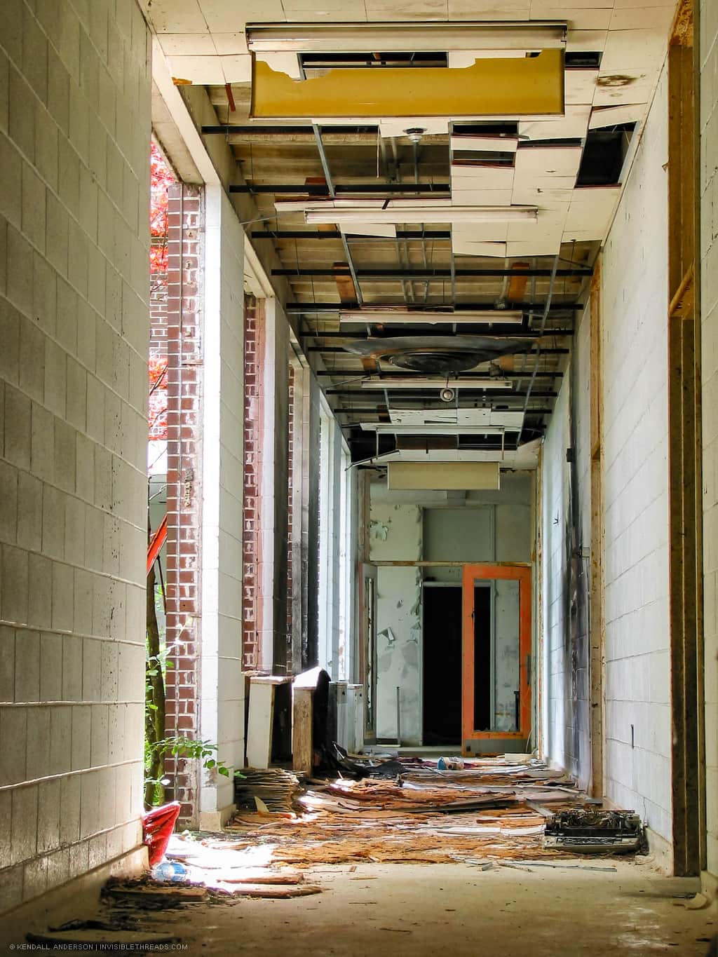 A long hallway filled with debris is brightly lit. All windows to an interior courtyard are broken, and the exit doors are missing all their glass.