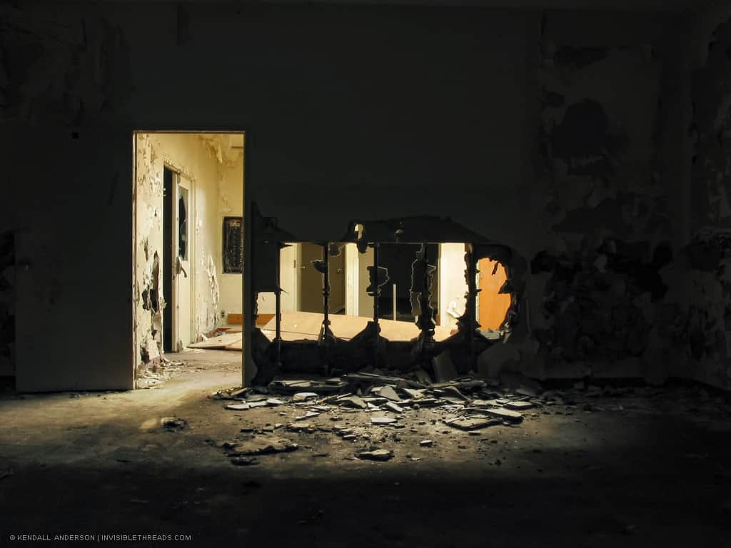 A door leads out of a dark room. The wall has 4 large holes in it from vandalism, exposing the wood studs in the wall. Debris from the holes is littered on the ground.