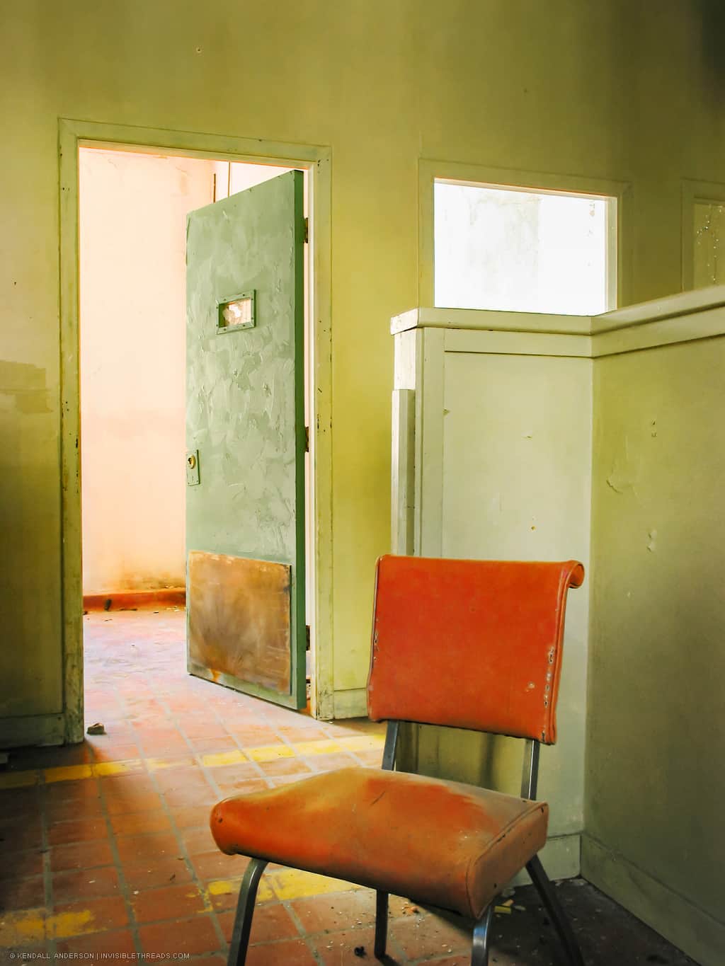An orange chair sits in front of an open green door. The door is solid, but has a very small slot window at eye level for viewing the occupants of the room. The floor is orange tile, dirty and dusty.