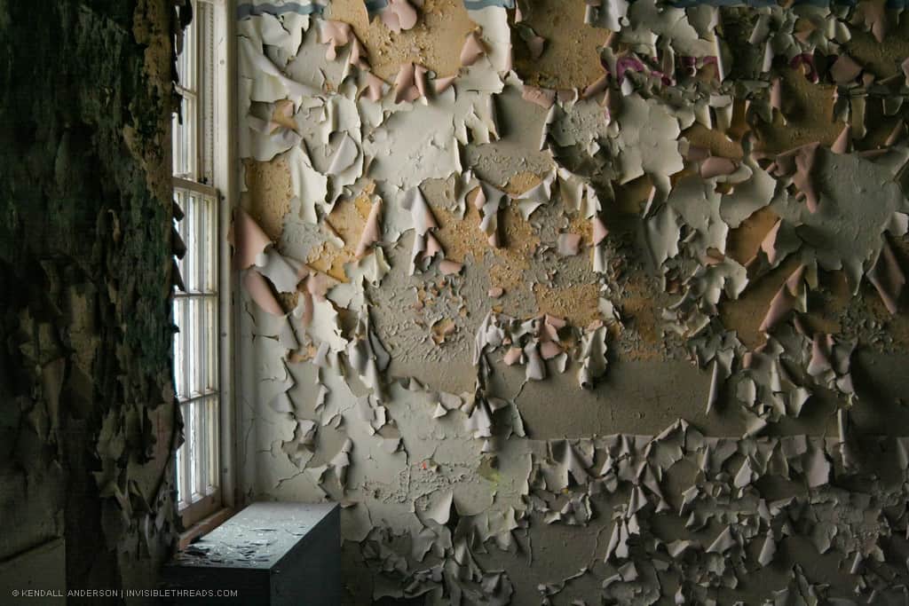 A window allows sunlight into the corner of a room. Layers of paint on the walls are peeling off in large pieces. The entire wall is peeling paint.