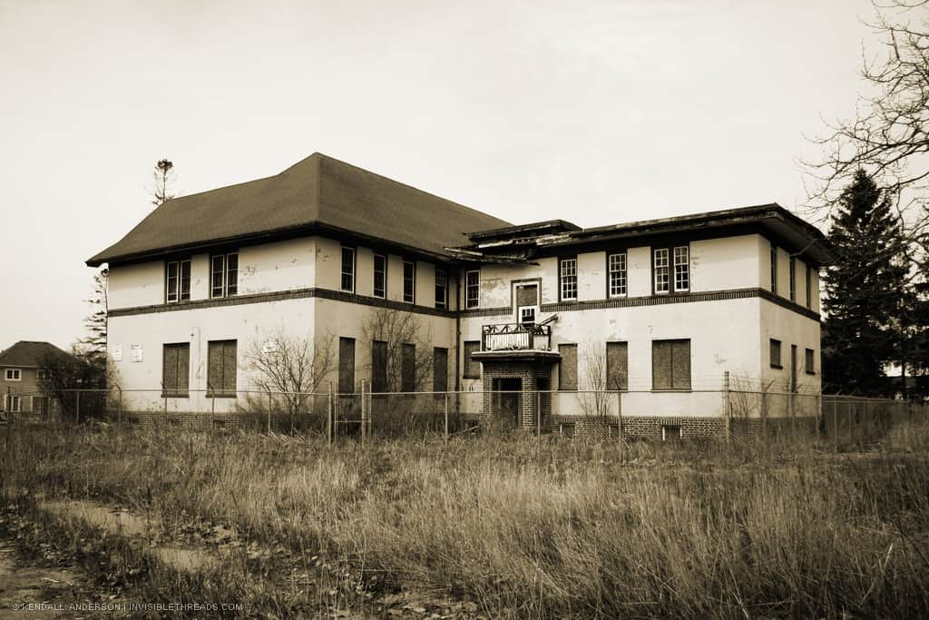 A chain-link fence surrounds the exterior of an abandoned 2-storey building. The grass is overgrown all around it.