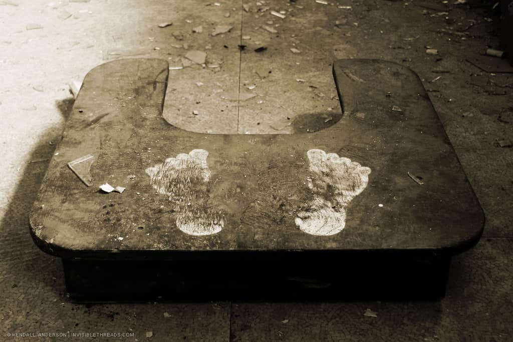 A small wooden platform with 2 footprints sits on a dirty floor.