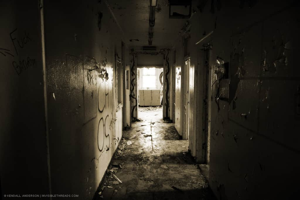 A dark hallway strewn with debris and walls covered in graffiti leads to a room with a window.