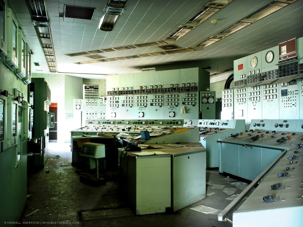 Multiple banks of instrument panels with gauges, switches, levers, dials and knobs are in a small room. The room is lit from the left with bright sunlight from a window. The control room is abandoned, and debris and papers are strewn about the desks and floor.