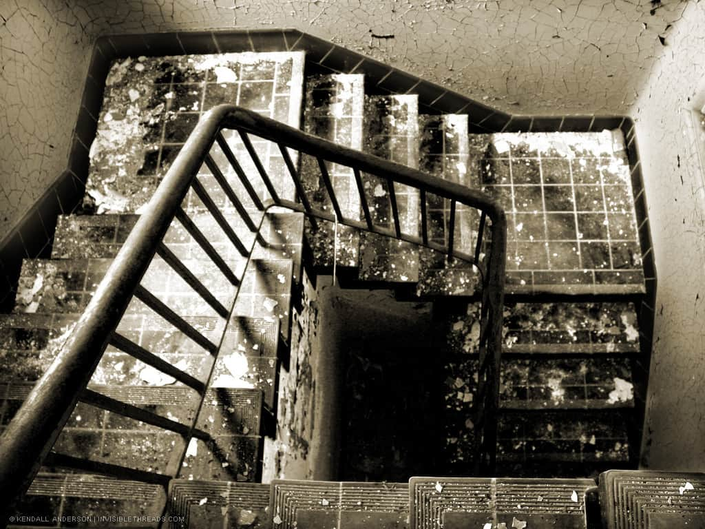 A spiral staircase is seen from above, descending. The stairs are covered in debris and paint chips, from the peeling paint on the cracked walls.