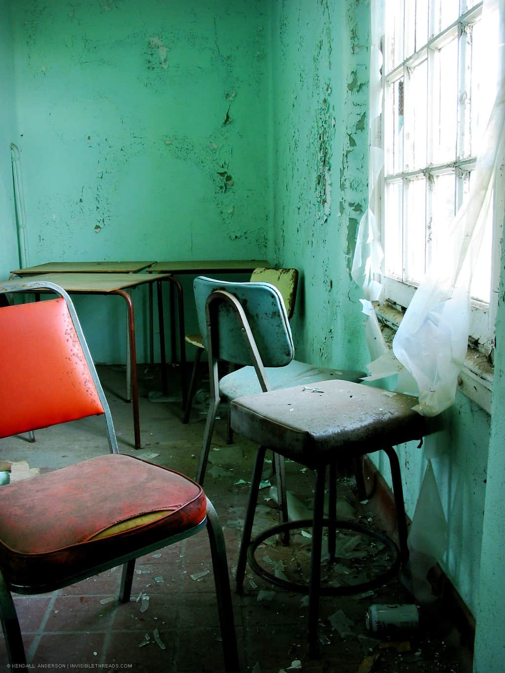 A green room with cracked walls is brightly lit from a large window. Inside the room are a few green chairs matching the wall colour, and a single bright orange chair.