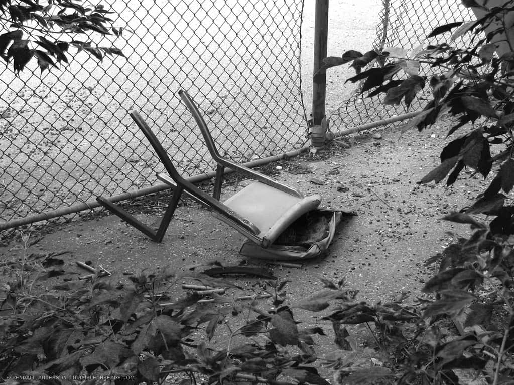 A broken chair is overturned, lying on the ground in front of a chain link fence with a gap in it.