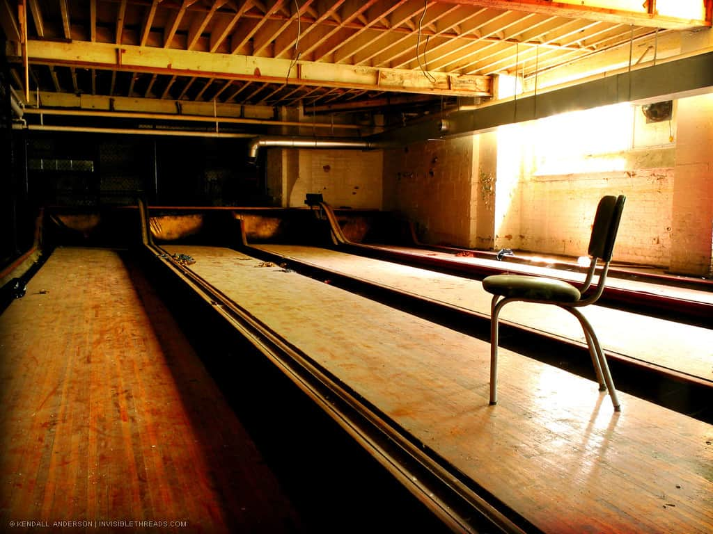 A single chair is placed in the middle of a bowling alley lane. The bowling alley is in the basement of a building, and an orange glow covers everything from the sunlight coming through the basement windows. The bowling lanes are dirty and covered in dust.