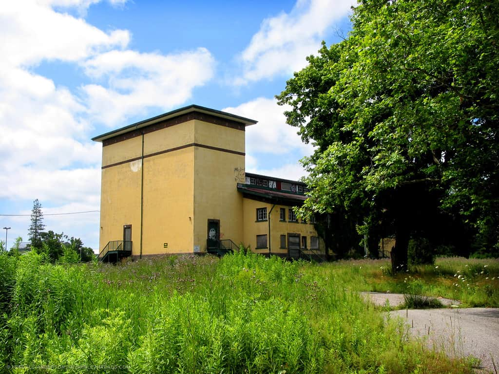 A recreation hall building with yellow exterior walls is surrounded by overgrown vegetation.