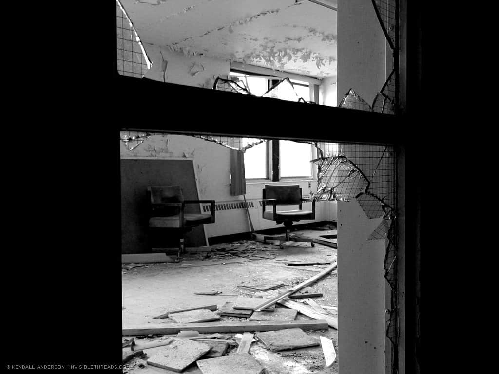 A room filled with debris and rubble is seen through the broken glass window in the wall.