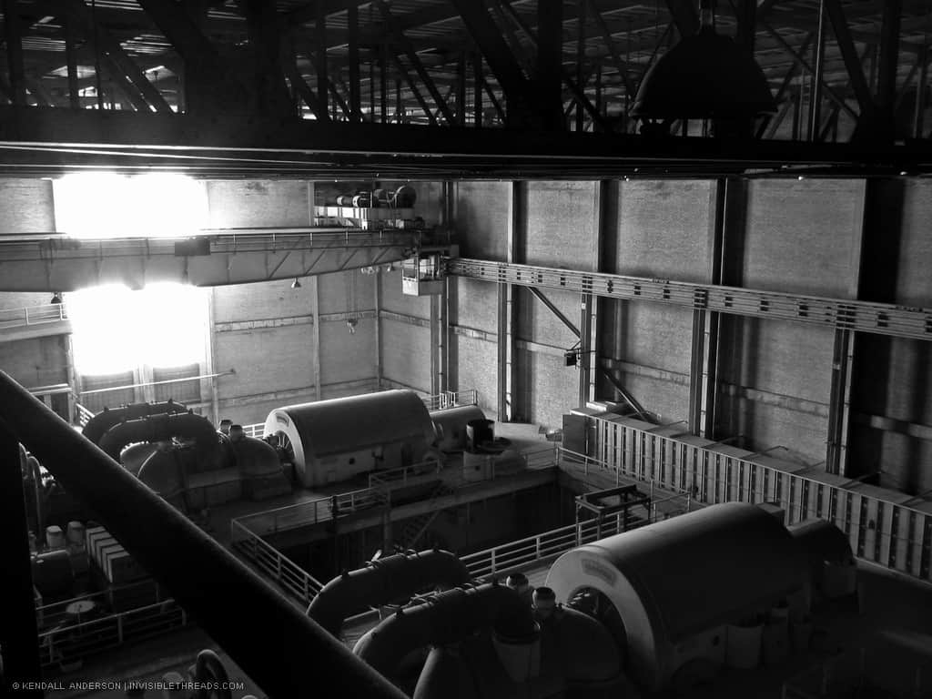 Two turbines/generating units rest on the main turbine floor of a generating station. The large beam of a crane on rail tracks is stationary, above the first turbine unit. Light enters the main hall from a large window to the left and above the turbines.