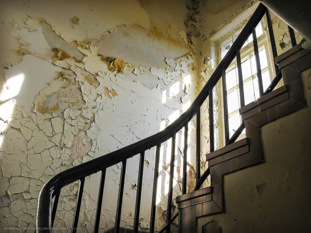 A railing leads up a spiral staircase. A window lets in light, highlighting the cracking and peeling paint on the walls.