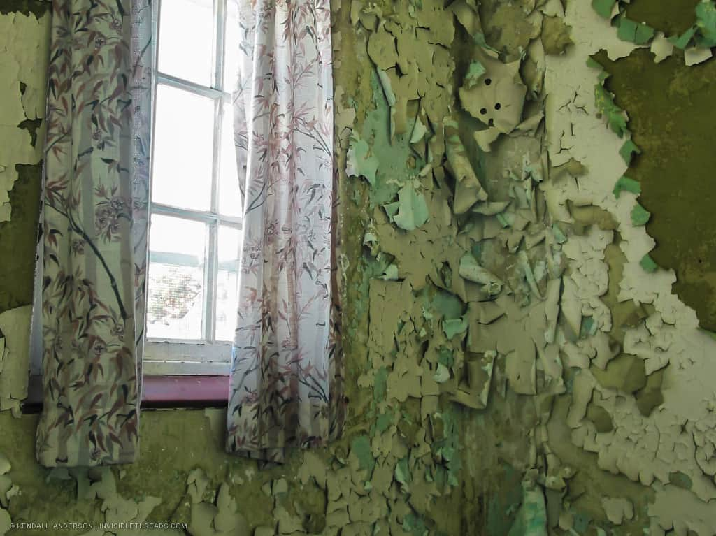 A window with pink curtains lets light into a room with severely peeling green paint. The walls are very decayed.