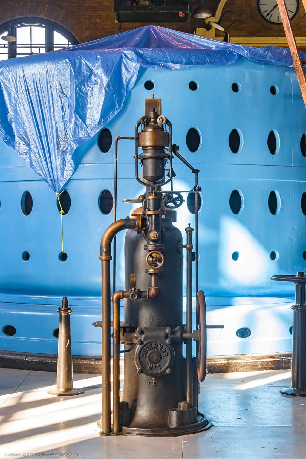 A 100 year-old flyball governor stands in front of a blue steel generator housing. The governor is fitted with polished brass pipes and valve wheels.