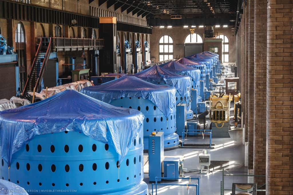 A series of 9 blue turbine generator heads are on the main floor of the power station. In the distance a large stair rises to a balcony leading to the control room. All of the generator heads are covered in plastic tarps.