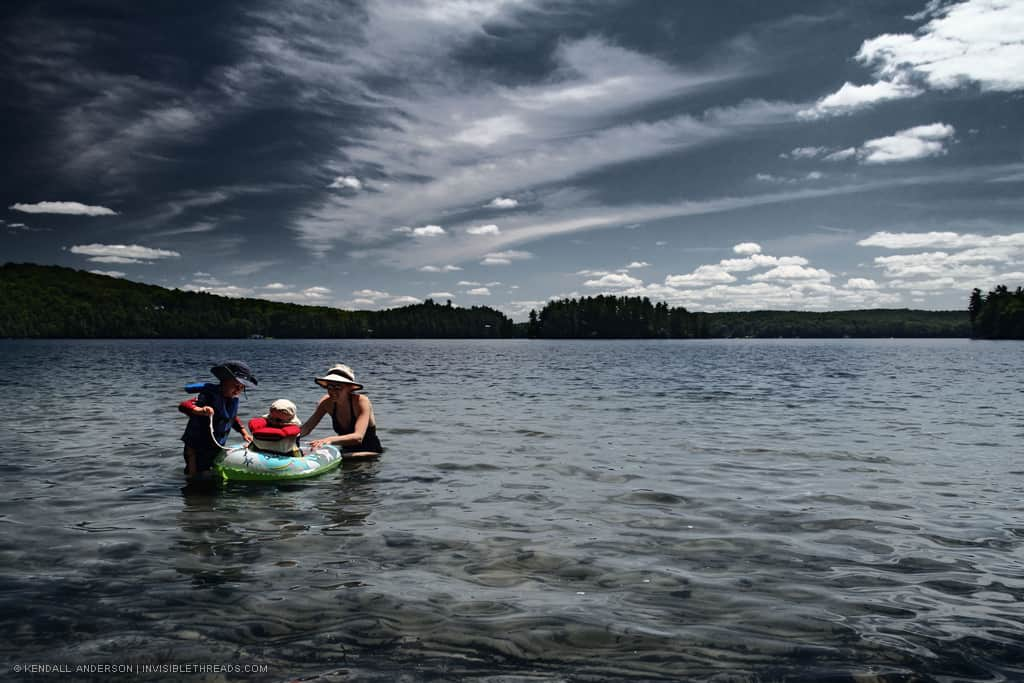 A woman with 2 kids is in the water. One child is in a small inflatable boat. The sky is dark and cloudy.