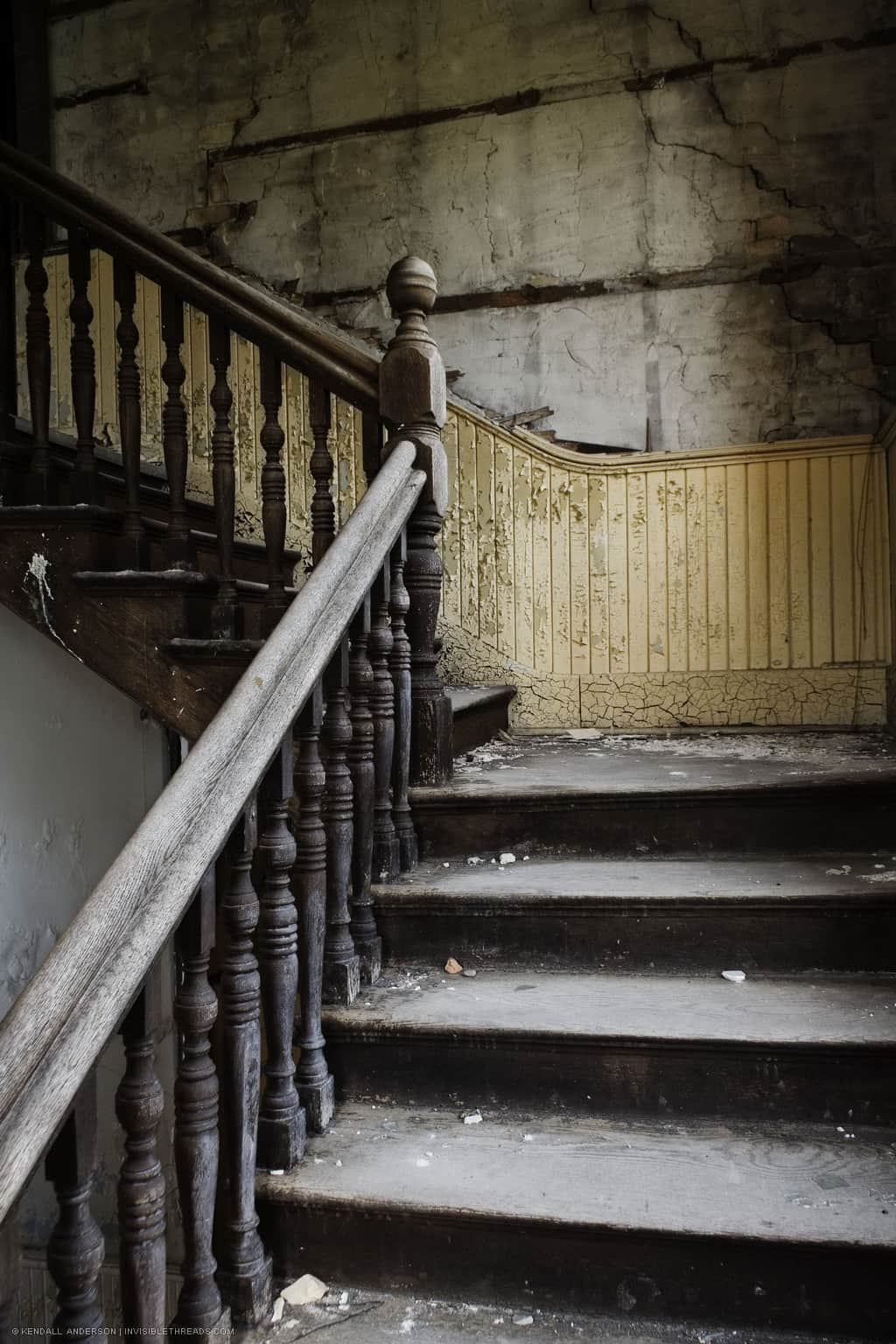 A wooden staircase ascends to the next floor. The paint is peeling on the walls and rubble and plaster from the walls lies on the stairs. A wooden handrail and banister rises up with the stairs.