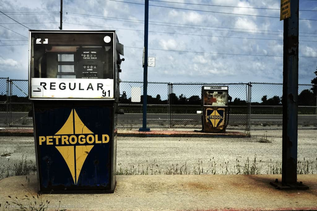 Two old gas pumps sit on concrete pads at a service station that is slowly being overgrown by grass. A chain link fence separates the station from the road beyond.