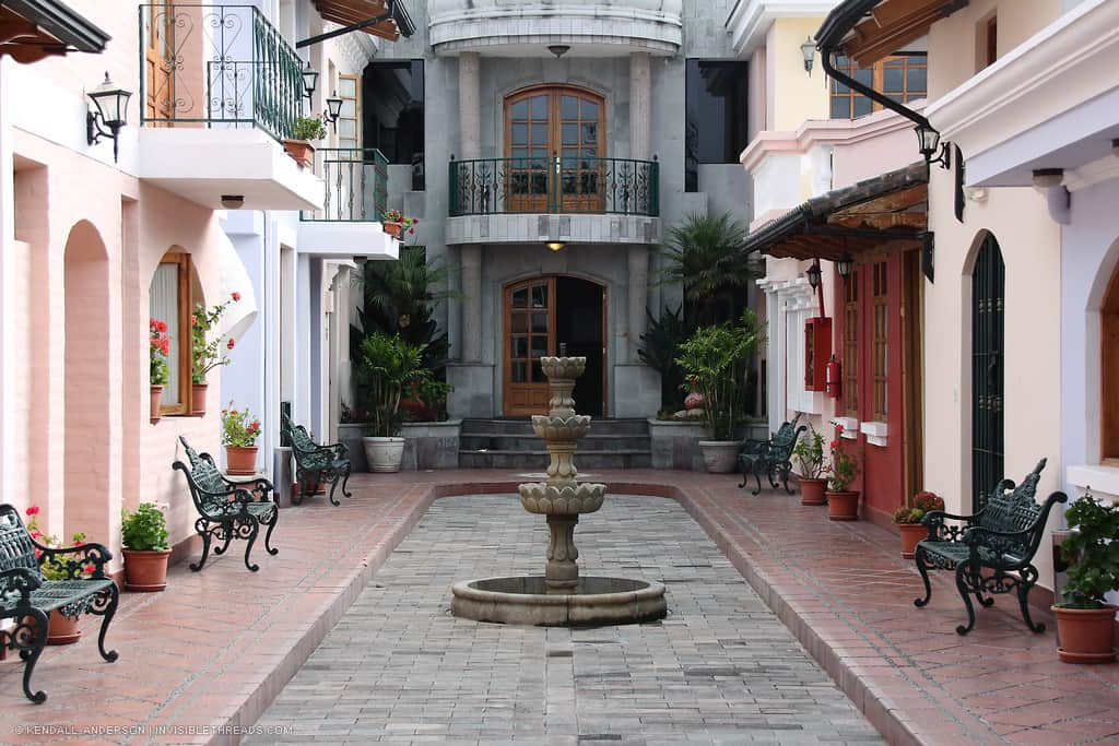 Hotel interior courtyard with stone fountain in center. Benches line the pastel walls, and a balcony overlooks the cobblestone ground below.